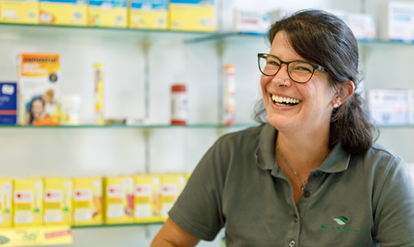 Apothekerin der Martinus Apotheke steht lachend vor dem Medikamentenregal in der Apotheke.